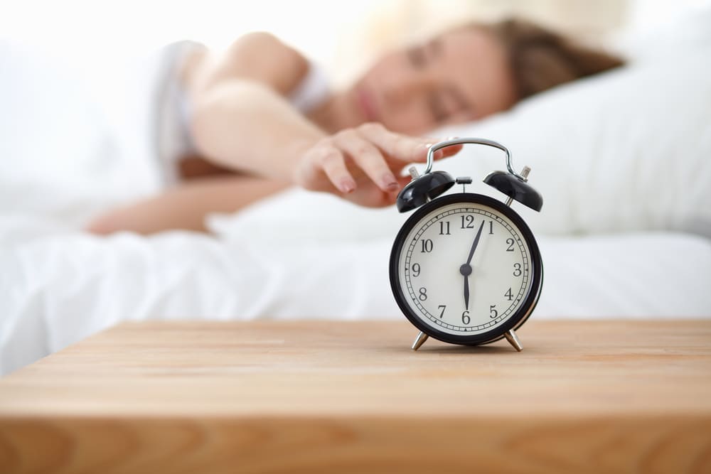 A woman in bed with her eyes closed, reaches to snooze the alarm clock on the table