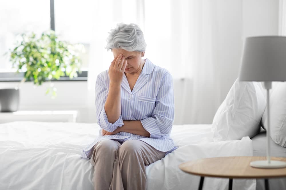 Older woman is sitting on a bed and pinching the bridge of her nose in pain